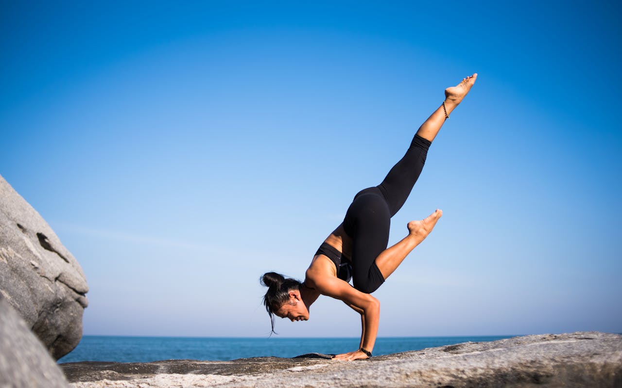 team-01 Woman performing a yoga pose on a rocky beach with ocean view and clear blue sky.