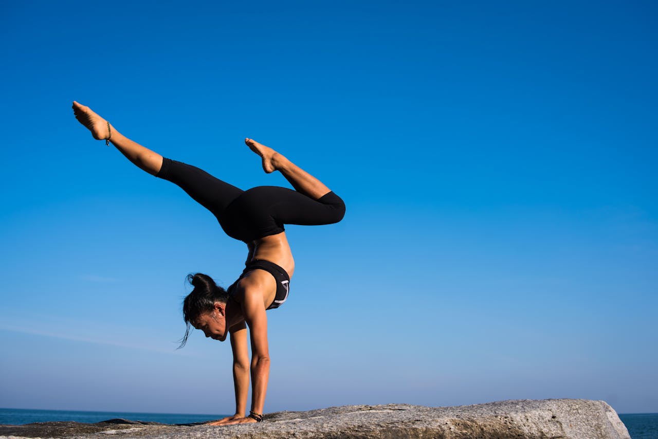 services-02 A woman performing a yoga handstand on a rock by the sea under a clear blue sky.